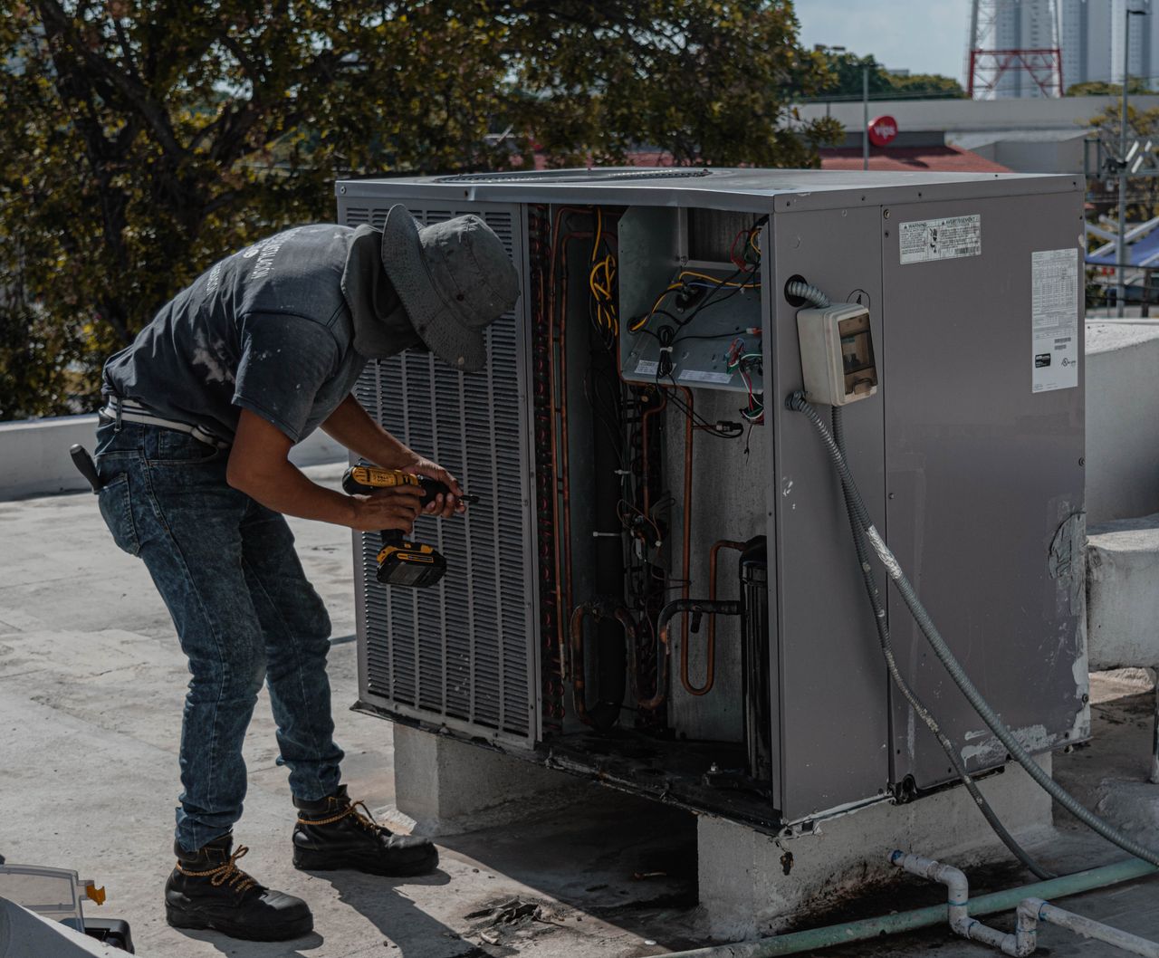 A worker in a bucket hat repairs an outdoor air conditioning unit on a rooftop.